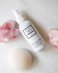A white bottle of Só Tonic Apple Cider Vinegar facial toning spray, styled on a white marble surface with pink crystal rocks and a cream coloured konjac sponge.