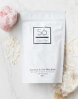 Só Luxury Coconut and Oat Milk Bath 300g pouch with scattered product around it and a pink crystal on the top left and a beige crystal on the bottom right