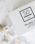 A white rectangular box of Só Sitz Bath Tea with black text, photographed on a white marble surface with a few quartz crystals nearby.