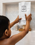 A child in a bathroom stands in front of a built-in shelf, holding a white pouch of Só Coconut & Oak Bath. The scene includes a white tiled wall, a glass jar of bath salts, and another Só product container on the shelf.