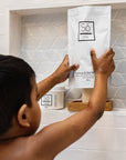 A child in a bathroom stands in front of a built-in shelf, holding a white pouch of Só Coconut & Oak Bath. The scene includes a white tiled wall, a glass jar of bath salts, and another Só product container on the shelf.