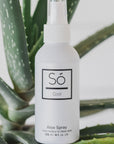 A white spray bottle of Só Cool Aloe Spray standing upright in front of green aloe vera leaves against a clean white background.