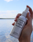 Hand holding a bottle of 'Sö Cool' aloe spray with a scenic background water and sky