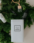 A styled flat-lay of Só luxury products arranged on a bed of green pine branches. The set includes a white spray hand sanitizer bottle, a black lip balm, a black bottle of sunlight essential oil blend, a clear glass perfume bottle with a beige cap, and a white rectangular tin with the Só logo.