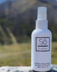 A white bottle of Só Outdoorsy outdoor body spray standing on a gray stone surface with a blurred mountain landscape in the background.