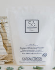 A package of Só Clean Oxygen Whitening Powder with a small scoop and a cloth beside it, on a white background with eucalyptus leaves.