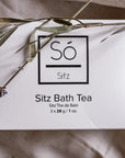A white rectangular box of Só Sitz Bath Tea with black text, photographed on a fabric surface with natural grass elements.