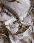 3 large tea bags styled on a linen coloured fabric one of the tea bags is sliced open showing the contents being various herbs including lavender with natural laveneder and styling elements.
