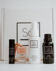 Collection of products with 'Só branding including a Lippy lip balm, small glass essential oil diffuser with a wood lid ands beige string, small peppermint hand sanitizer and a black bottle of Sunlight essential oil blend on a light background with a Só branded gift box in the background.