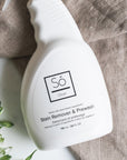 White spray bottle branded with 'Só Clean Stain Remover + Prewash' on a light marble background with light brown fabric and floral greenery.