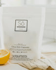 A white package of Só Clean Citrus Dish Capsules with a half lemon beside it, indicating a citrus scent on a blurred light background with a beige fabric.