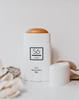 A white plastic deodorant style container branded Só Salve Fire Salve a warming muscle salve in 50 g twist-up stick styled on a white backdrop with the cap off showing an orange product and natural floral elements.