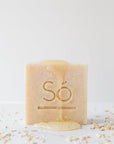 A beige bar of soap branded Só standing upright with honey dripping down the front and oats sprinkled around on a white background.