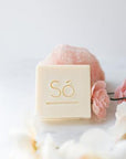 A cream coloured square bar of soap branded with Só styled on a light coloured background with a rose quartz stone and faux florals.