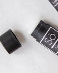 A stick of lip balm with the brand name Só Lippy placed on a surface, open, with its cap next to it, against a marble background.