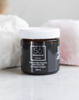 A amber glass jar with white lid of Só Silver Mission Falls Premium Ionic Silver Gel, 100 ml, set on a marble surface with white and pink rocks in the background.