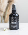A black spray bottle of Só Silver Mission Falls Premium Ionic Silver Solution Topical Spray with a clear cap, standing on a white marble surface with a small white flower and blurred dried pampas grass in the background.