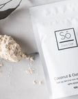 A package of Só Luxury Coco Oat Milk Bath powder with some scattered on a gold spoon beside it, all placed on a white marble surface.