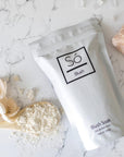 A white stand-up pouch of Só Blush Blush Soak set on a white marble surface with scattered powder, a spoonful of the powder and a decorative pink stone.