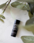 A black lip balm stick with a white logo and text, labeled 'Só Lippy Natural Lip Balm', placed on a light marble surface next to green eucalyptus leaves.