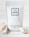 A package of Só Luxury Coco Oat Milk Bath powder with some scattered on a gold spoon beside it, all placed on a white marble surface.