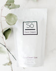 A packaged coconut milk bath soak, labeled 'Só Luxury Coco Cream Soak' on a marble surface with a leafy green plant stem.