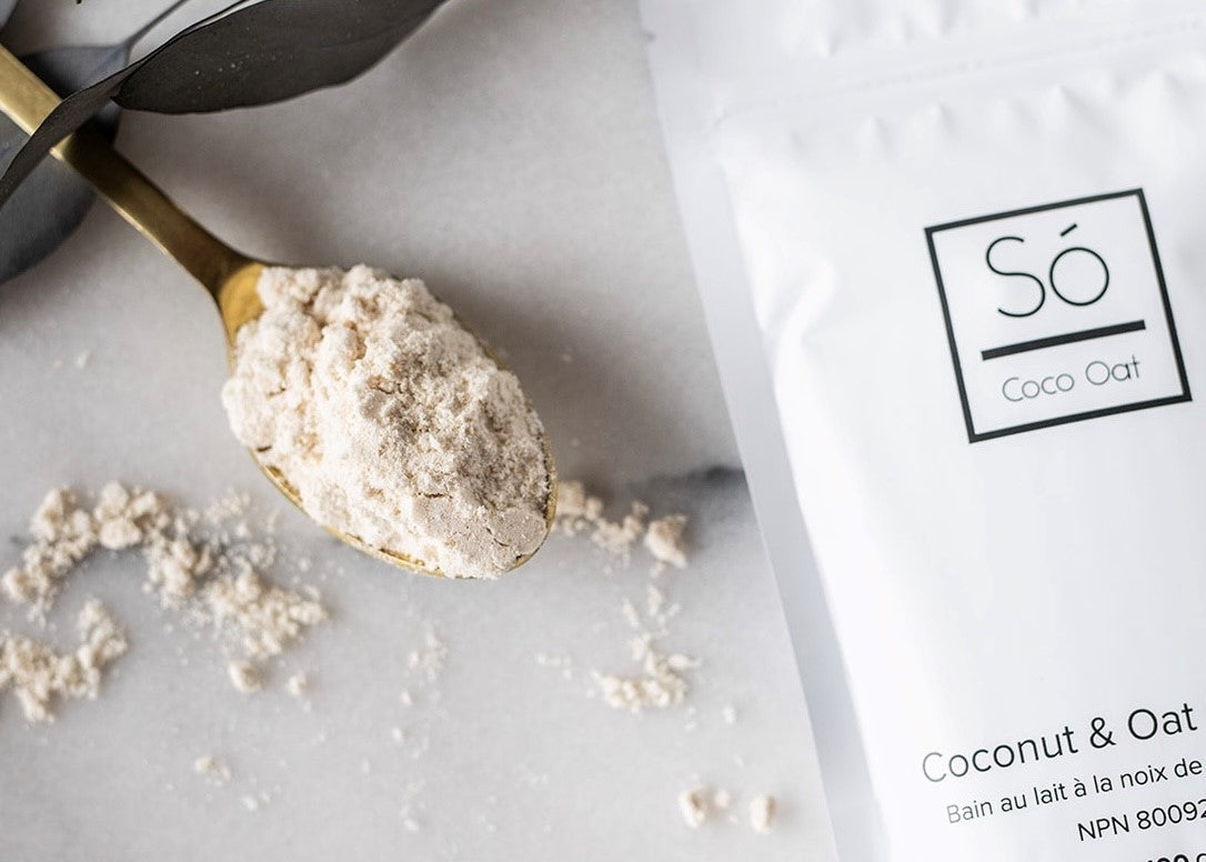 A white bag of Só Coco Oat Milk Bath styled on a marble background with a golden coloured spoon with beige powder on the spoon and black floral elements.