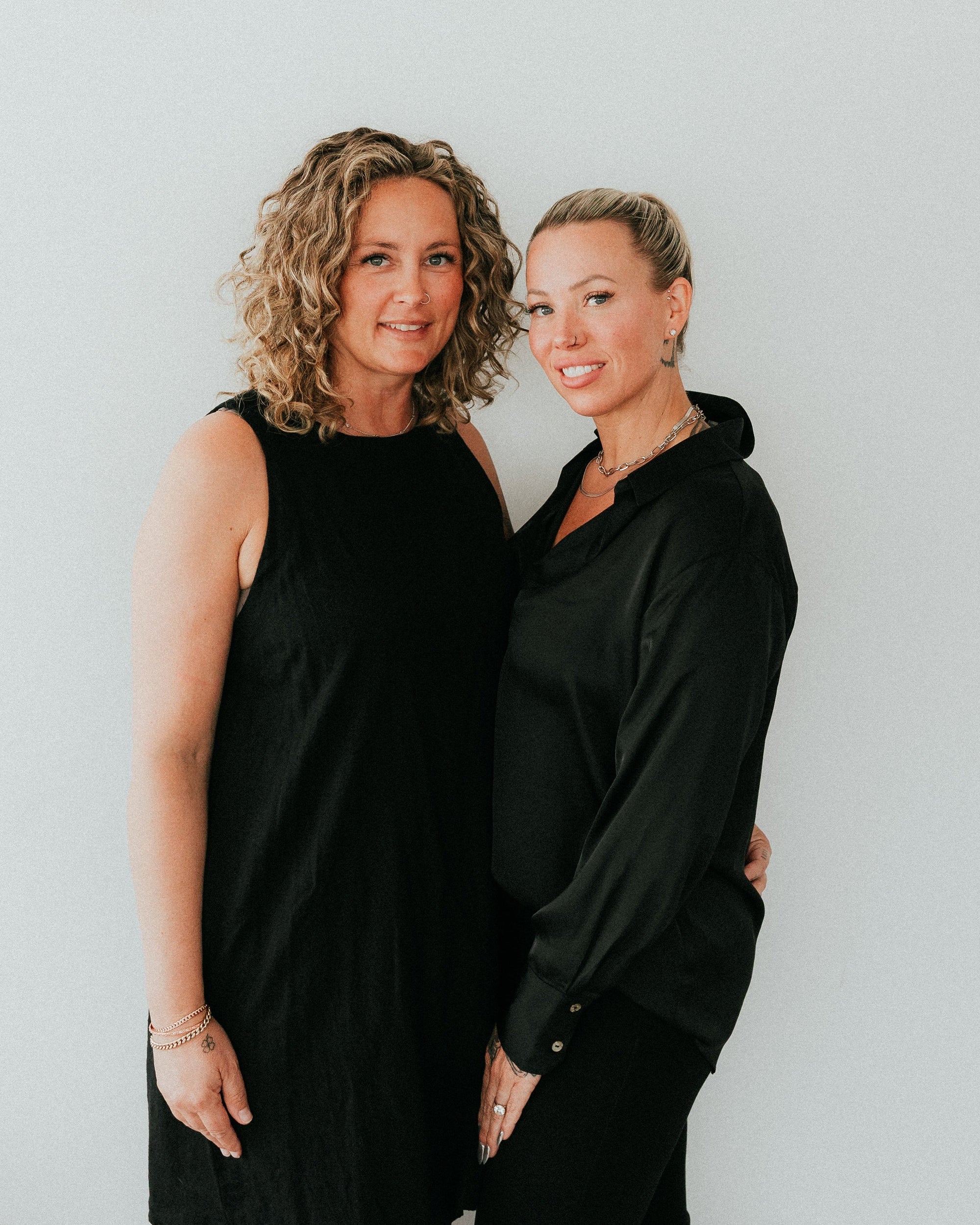 Two women standing side-by-side against a plain white wall, both wearing black. The woman on the left has curly blonde hair and wears a sleeveless black dress, while the woman on the right has short blonde hair and wears a black long-sleeve button-up shirt and black pants.