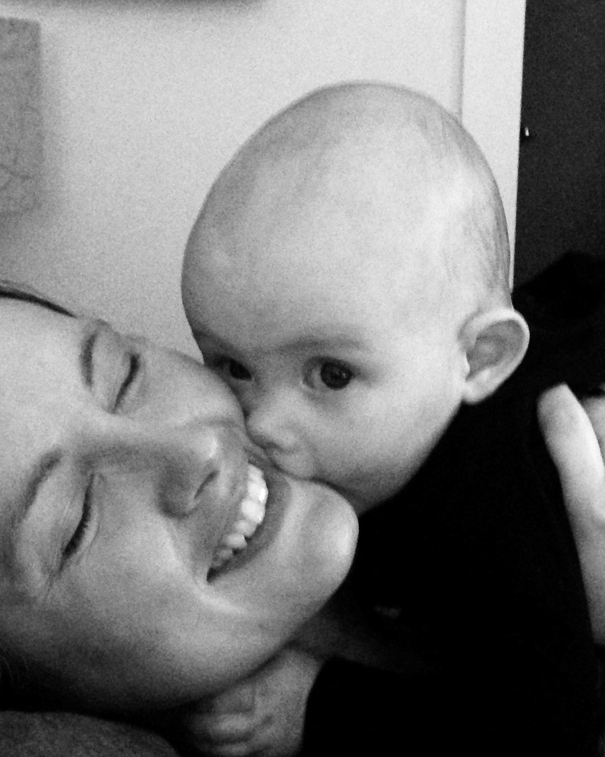 Black and white photo of a woman and a baby kissing mom's face mom's eyes are closed and she is smiling and the baby is looking into the camera.