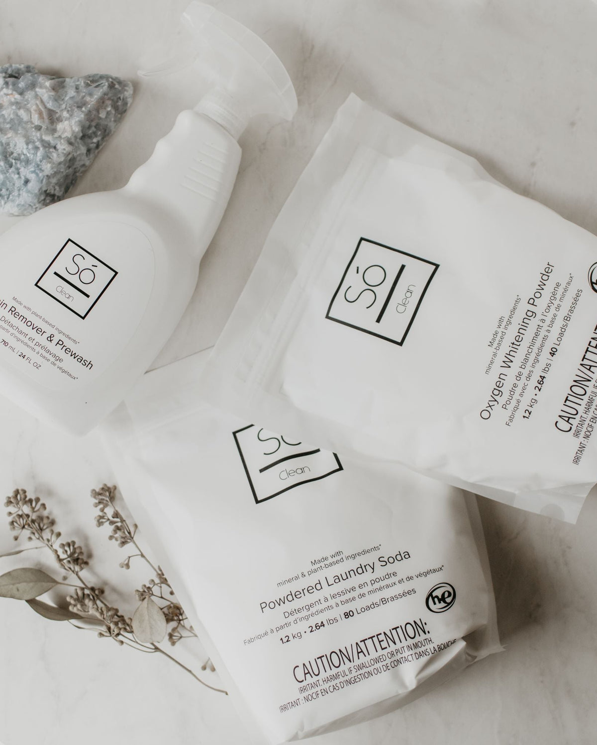 Three white Só Clean laundry and stain care products arranged on a light marble surface: a stain remover spray bottle, a powdered laundry soda bag, and a whitening powder bag, with decorative stones and dried flowers nearby.