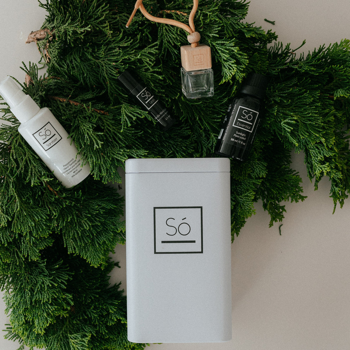 A styled flat-lay of Só luxury products arranged on a bed of green pine branches. The set includes a white spray hand sanitizer bottle, a black lip balm, a black bottle of sunlight essential oil blend, a clear glass perfume bottle with a beige cap, and a white rectangular tin with the Só logo.