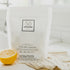 A white package of Só Clean Citrus Dish Capsules with a half lemon beside it, indicating a citrus scent on a blurred light background with a beige fabric.