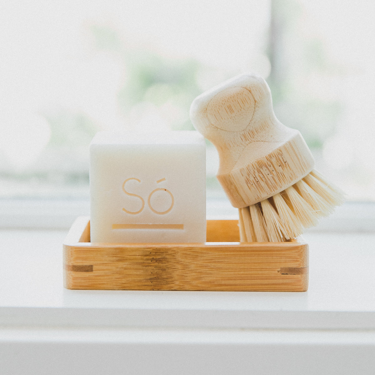 A bamboo soap dish holding a white bar of soap embossed with the brand 'Só' and a small wooden-handled cleaning brush with natural bristles, set on a bright white surface near a softly blurred window.