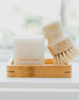 A bamboo soap dish holding a white bar of soap embossed with the brand 'Só' and a small wooden-handled cleaning brush with natural bristles, set on a bright white surface near a softly blurred window.