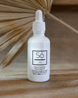 A white bottle with a dropper cap labeled 'Só Oil' with a scent described as Island Coconut. It is placed on a wooden surface with a dried palm leaf in the background.