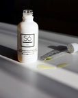 A white bottle with a dropper containing unscented oil, placed on a surface with a few drops of oil visible in front of the bottle.