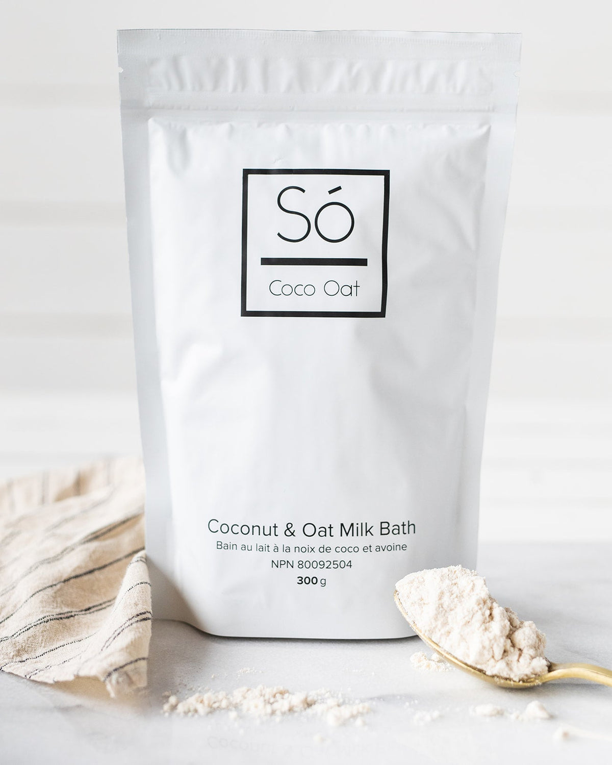 A package of Só Luxury Coco Oat Milk Bath powder with some scattered on a gold spoon beside it, all placed on a white marble surface.